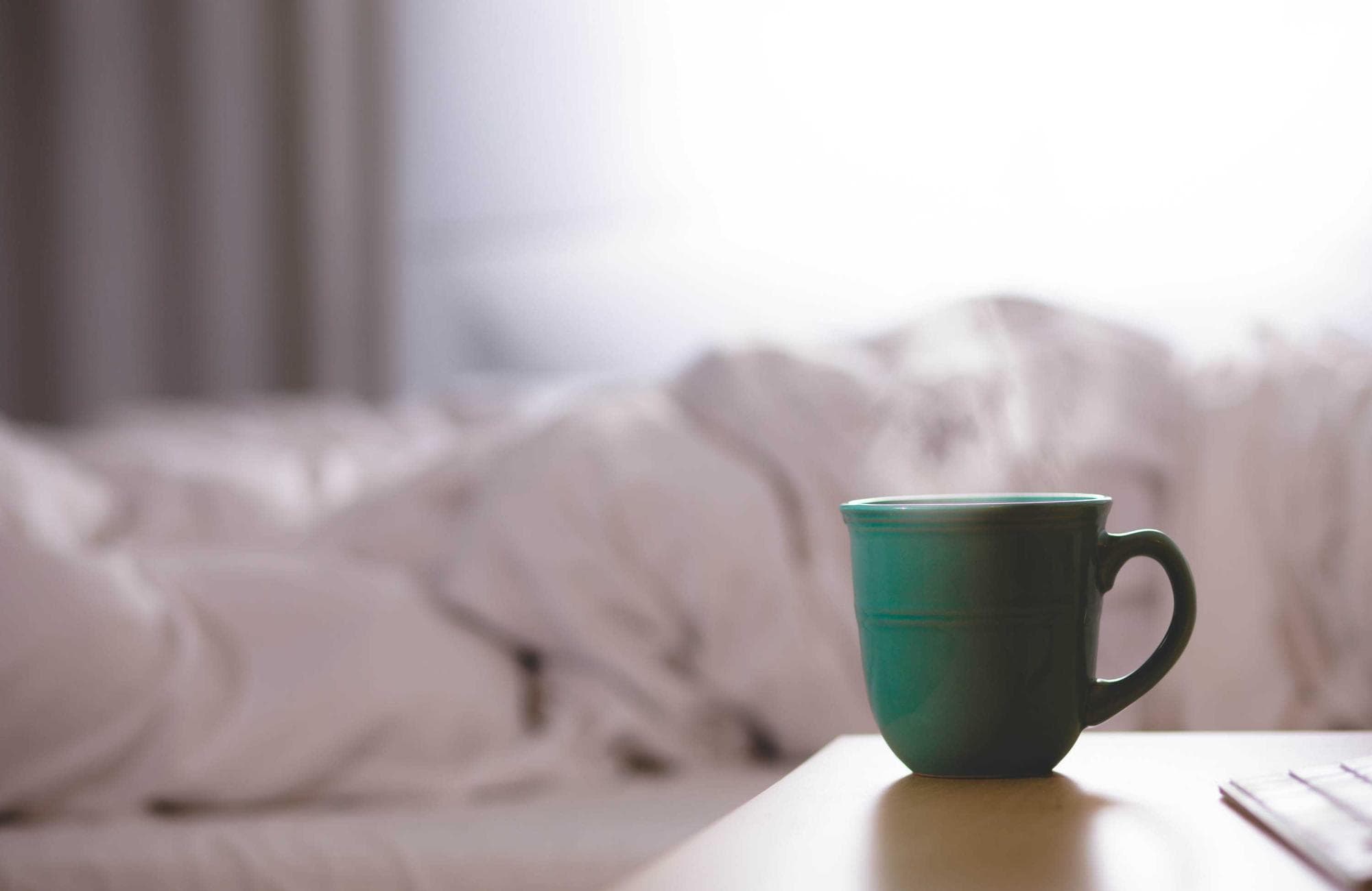 A mug on a bedside table beside soft white bedding in morning light